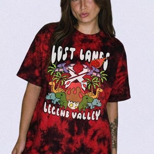 May include: A red and black tie-dye t-shirt featuring a graphic print of dinosaurs, palm trees, and a volcano. The text "LOST LANDS" appears at the top and "LEGEND VALLEY" at the bottom in white lettering, with a central white X symbol.