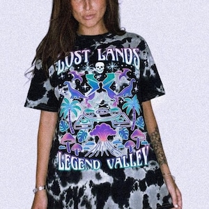 May include: A black and white tie-dye t-shirt featuring a graphic print with the text LOST LANDS and LEGEND VALLEY. The design includes a central figure in a meditative pose, a skull, palm trees, mushrooms, and a volcano in shades of blue and purple.