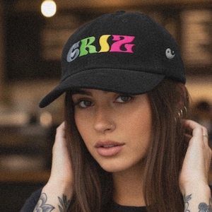 May include: Black baseball cap with the word "GRSZ" in colorful lettering. The cap has a curved brim and a small emblem on the side. The person in the image is wearing a black shirt.