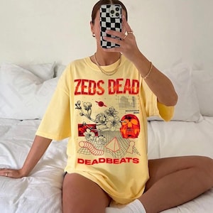 Puede incluir: Una camiseta de manga corta de color amarillo pálido con un estampado gráfico que incluye el texto ZEDS DEAD y DEADBEATS. El diseño presenta una calavera roja, ramas de cerezo, un patrón de cuadrícula, una pirámide y un pequeño planeta en tinta roja y negra.
