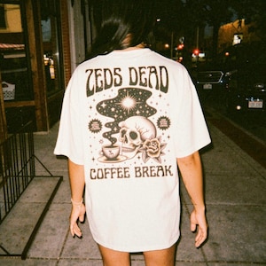 Puede incluir: Camiseta color crema con un gráfico en la espalda que presenta una calavera, una taza de café, una rosa y las palabras "ZEDS DEAD" y "COFFEE BREAK" en un estilo vintage. La camiseta la lleva una persona caminando por una acera.