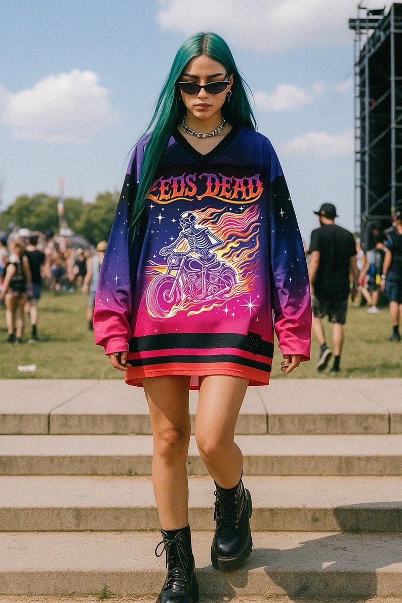 May include: A long-sleeved jersey-style shirt with a vibrant design. The shirt features a skeleton riding a motorcycle engulfed in flames, with the text "Zeds Dead" across the chest. The color scheme transitions from purple to pink.