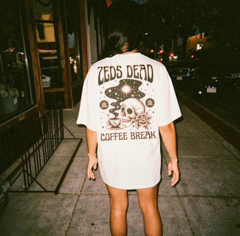 May include: Cream-colored t-shirt with a graphic on the back featuring a skull, coffee cup, rose, and the words "ZEDS DEAD" and "COFFEE BREAK" in a vintage style. The shirt is worn by a person walking on a sidewalk.