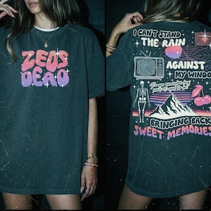 May include: Dark green t-shirt with the text "ZEUS DEAD" in pink, orange, and purple. The back of the shirt features a graphic with the text "I CAN'T STAND THE RAIN AGAINST MY WINDOW" and a skeleton, mountains, and cherries.