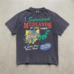 May include: A charcoal gray short-sleeve t-shirt featuring a graphic print with the text I Survived MUDLANDS AT LOST LANDS, LEGEND VALLEY OHIO, A Little Dirt Never Hurt, and 2026. The design includes a green dinosaur and a yellow map shape.