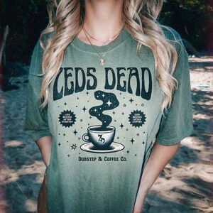 May include: Olive green t-shirt with the text "CEDS DEAD" in a retro font. Below the text is a graphic of a coffee cup with a swirling design and the text "Dubstep & Coffee Co."