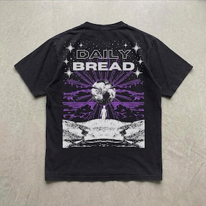Pu&ograve; includere: Una t-shirt nera a maniche corte con una stampa grafica sul retro. Il design mostra il testo DAILY BREAD sopra una grande luna, la sagoma di una persona in un paesaggio roccioso, raggi di luce viola e stelle bianche.
