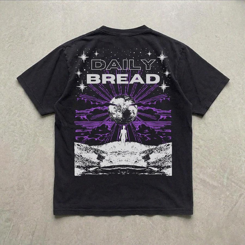 Pu&ograve; includere: Una t-shirt nera a maniche corte con una stampa grafica sul retro. Il design mostra il testo DAILY BREAD sopra una grande luna, la sagoma di una persona in un paesaggio roccioso, raggi di luce viola e stelle bianche.