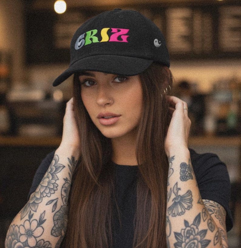 May include: Black baseball cap with the word "GRSZ" in colorful lettering. The cap has a curved brim and a small emblem on the side. The person in the image is wearing a black shirt.