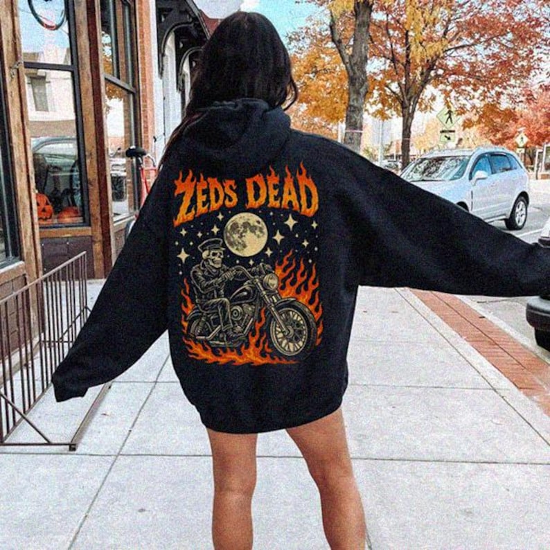May include: Black hooded sweatshirt with the text "Zeds Dead" in orange. The back graphic features a skeleton riding a motorcycle surrounded by flames, a full moon, and stars. The sweatshirt is worn outdoors.