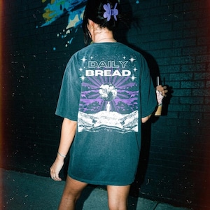Puede incluir: Camiseta holgada de color verde azulado oscuro con las palabras "DAILY BREAD" sobre un gráfico de un paisaje morado y blanco con un sol y una figura. La persona lleva una pinza para el pelo morada.