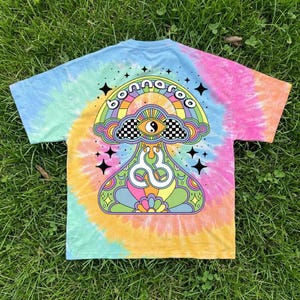 May include: A short-sleeved tie-dye t-shirt featuring a colourful graphic on the back. The design includes a mushroom shape, a yin-yang eye, a rainbow, and the text "bonnaroo" in black letters, surrounded by small black star icons.