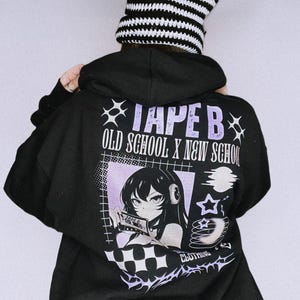 May include: Black hoodie with a graphic on the back featuring an anime-style character holding a tape. The text on the hoodie reads "TAPE B OLD SCHOOL X NEW SCHOOL CLOTHING." A black and white striped cat ear hat is worn.
