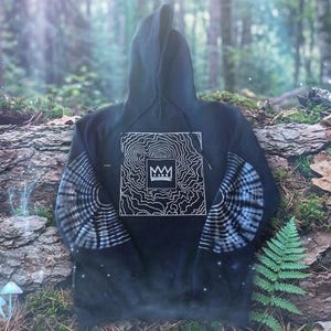 May include: A black hooded sweatshirt featuring a white graphic print on the chest of a crown inside a square surrounded by topographic lines. The sleeves display a circular tie-dye pattern in shades of gray and white, with a front kangaroo pocket.