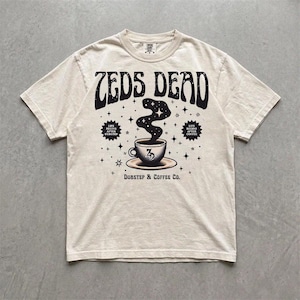 Puede incluir: Una camiseta de manga corta color crema con un gráfico negro de una taza de café con vapor elevándose hacia un cielo estrellado. El texto dice ZEDS DEAD, LIFE BEGINS AFTER DUBSTEP, LIFE BEGINS AFTER COFFEE y DUBSTEP & COFFEE CO.