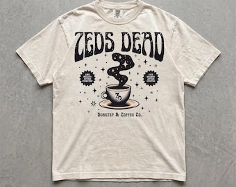 Può includere: Una t-shirt a maniche corte color crema con una grafica nera di una tazza di caffè con vapore che sale verso un cielo stellato. Il testo riporta ZEDS DEAD, LIFE BEGINS AFTER DUBSTEP, LIFE BEGINS AFTER COFFEE e DUBSTEP & COFFEE CO.