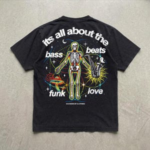 May include: A black t-shirt featuring a graphic of a skeleton holding a saxophone, surrounded by a mushroom, stars, and geometric lines. The text reads: its all about the bass beats funk love and SOUNDWAVE CLOTHING in white lettering.