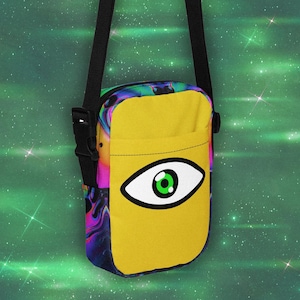 May include: A rectangular crossbody bag featuring a solid yellow front pocket with a large graphic of a stylised eye with a bright green iris. The sides and top of the bag display a vibrant, swirling psychedelic pattern in shades of purple, blue, and pink.