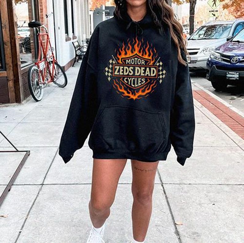 May include: Black hooded sweatshirt with a graphic design featuring the text "MOTOR ZEDS DEAD CYCLES" in a flame-like design. The sweatshirt has a front pocket and a drawstring hood. The design is in orange and yellow.