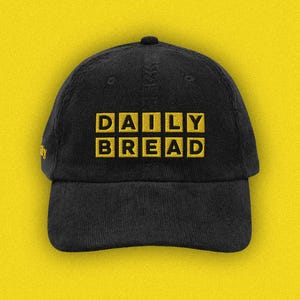Puede incluir: Una gorra de béisbol de pana negra con el texto DAILY BREAD bordado en letras amarillas en la parte frontal. La gorra cuenta con una corona estructurada, una visera curva y un pequeño detalle bordado en amarillo en el panel lateral.