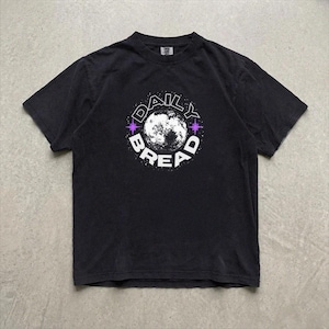 Pu&ograve; includere: Una t-shirt nera a maniche corte con una grafica circolare sul petto. Il design presenta una luna bianca al centro, circondata dalla scritta "DAILY BREAD" in lettere maiuscole bianche, accentuata da piccole stelle viola a quattro punte.