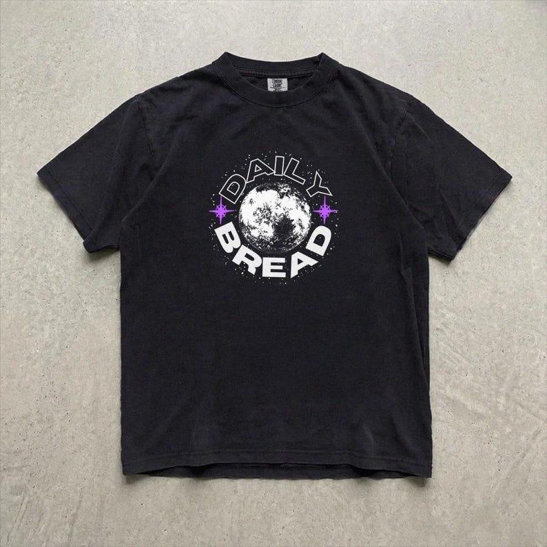 Pu&ograve; includere: Una t-shirt nera a maniche corte con una grafica circolare sul petto. Il design presenta una luna bianca al centro, circondata dalla scritta "DAILY BREAD" in lettere maiuscole bianche, accentuata da piccole stelle viola a quattro punte.