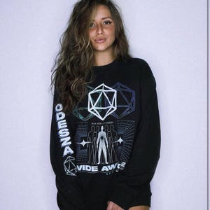 May include: A black long-sleeve sweatshirt featuring a white graphic print on the front and sleeve. The design includes a geometric icosahedron, stylized human figures, and the text ODESZA and WIDE AWAKE with the phrase HOW DID I GET HERE?