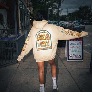 May include: A beige hoodie with the text "Freshly Baked Daily Bread" and a breakfast graphic on the back. The sleeves feature illustrations of eggs, bacon, and toast. The hoodie is worn outdoors.