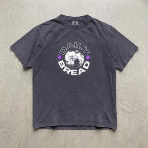 Pu&ograve; includere: Una t-shirt a maniche corte grigio antracite con una grafica circolare sul petto. Il design presenta un'illustrazione dettagliata della luna al centro, circondata dalla scritta DAILY BREAD in lettere maiuscole bianche e piccoli accenti a stella viola.