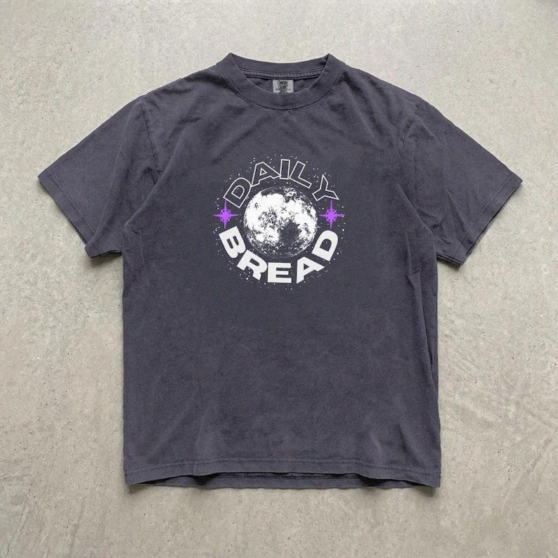 Pu&ograve; includere: Una t-shirt a maniche corte grigio antracite con una grafica circolare sul petto. Il design presenta un'illustrazione dettagliata della luna al centro, circondata dalla scritta DAILY BREAD in lettere maiuscole bianche e piccoli accenti a stella viola.