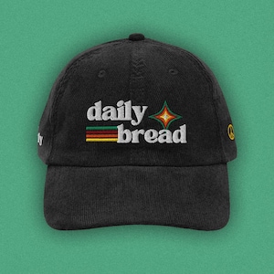 Peut inclure: Une casquette de baseball en velours côtelé noir avec le texte brodé en blanc daily bread. Le motif comprend trois bandes horizontales rouge, jaune et verte, une étoile à quatre branches et un petit signe de paix jaune sur le côté.