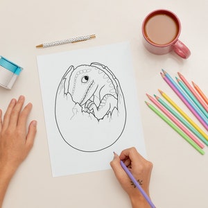 May include: A black and white line drawing of a dinosaur hatching from an egg. The drawing is on a white sheet of paper. A person is colouring the drawing with a purple coloured pencil.
