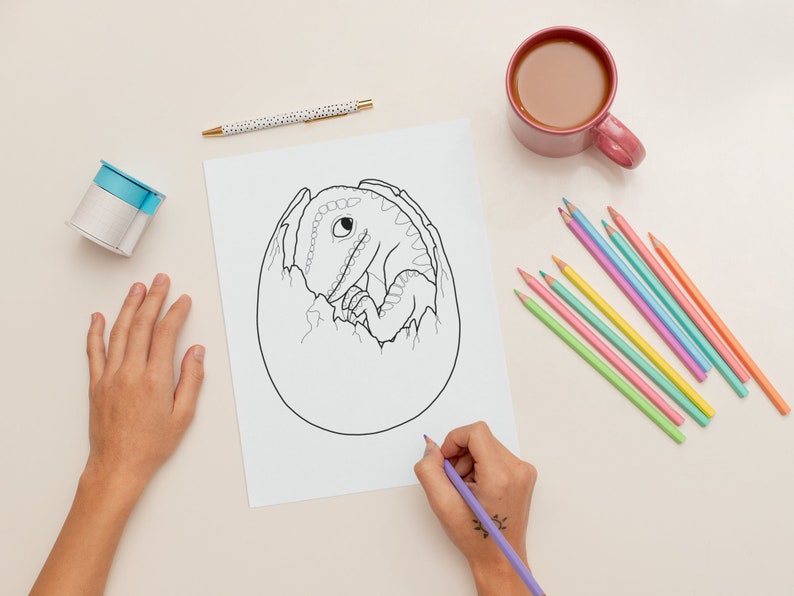 May include: A black and white line drawing of a dinosaur hatching from an egg. The drawing is on a white sheet of paper. A person is colouring the drawing with a purple coloured pencil.