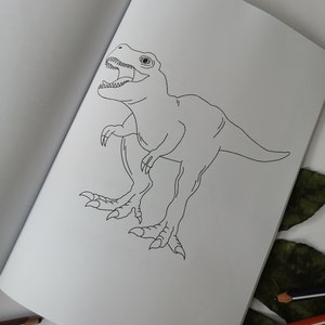 May include: An open coloring book featuring a detailed black and white illustration of a Tyrannosaurus Rex. The dinosaur is depicted with its mouth open, revealing teeth, and is positioned on a white page. Colored pencils are visible.