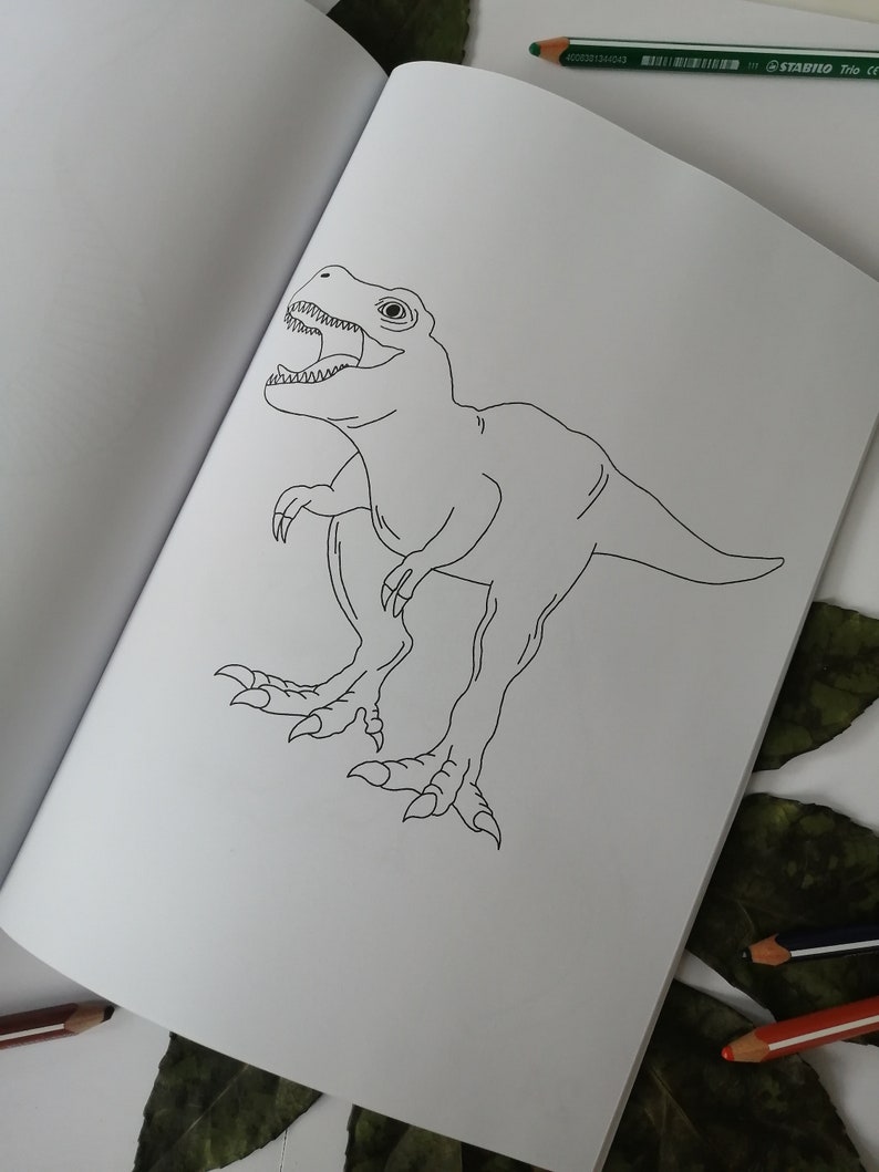 May include: An open coloring book featuring a detailed black and white illustration of a Tyrannosaurus Rex. The dinosaur is depicted with its mouth open, revealing teeth, and is positioned on a white page. Colored pencils are visible.