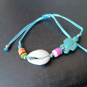 Può includere: Un braccialetto di corda blu con una conchiglia bianca, un ciondolo a forma di tartaruga turchese e perline colorate.