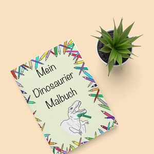 May include: A coloring book titled "Mein Dinosaurier Malbuch" with a dinosaur illustration, surrounded by a border of colorful pencils. A small potted succulent sits beside the book on a light beige surface.