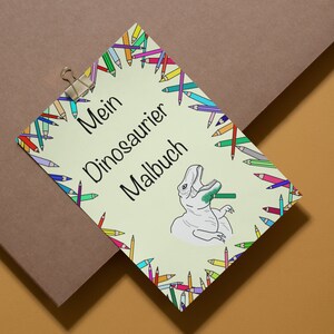 May include: A light green coloring book titled "Mein Dinosaurier Malbuch" with a cartoon dinosaur illustration. The book is surrounded by a border of colorful pencils and held by a silver clip.