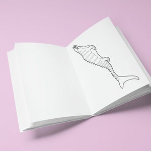 May include: An open colouring book with a black and white illustration of a fish on the right page. The book is on a pink surface.