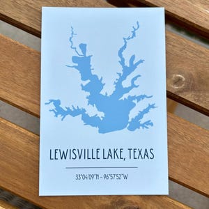 May include: A rectangular print featuring a light blue silhouette map of Lewisville Lake, Texas. Below the map, the text reads "LEWISVILLE LAKE, TEXAS" followed by a horizontal line and the coordinates "33°04'09"N - 96°57'52"W" in black font.