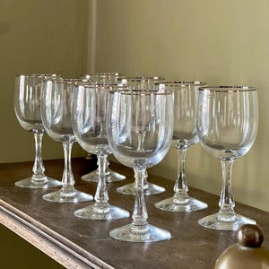 May include: A set of seven clear glass goblets arranged in rows. Each goblet features a smooth, rounded bowl, a slender stem with a decorative faceted base, and a thin metallic silver rim around the top edge of the glass opening.