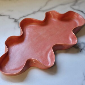Può includere: Un vassoio in ceramica color corallo, dalla forma libera e ondulata. Il vassoio ha una finitura liscia e lucida ed è esposto su una superficie marmorizzata. Questo vassoio decorativo è adatto per servire o esporre.