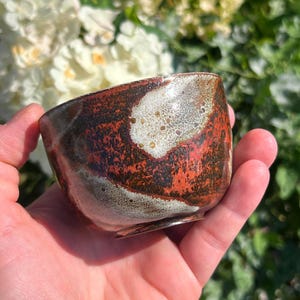 May include: A small, hand-held ceramic bowl featuring a mottled glaze with deep rust-red, dark brown, and creamy white patches. The surface has a textured, organic appearance with visible variations in color and a smooth, glossy finish throughout.