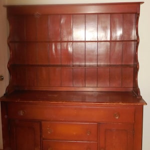 May include: A vintage wooden hutch with a red-brown finish. The hutch has a top shelf with two shelves below, and a bottom cabinet with three drawers and two doors.