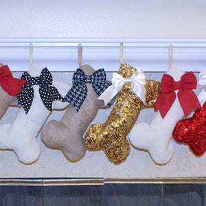May include: Nine bone-shaped Christmas stockings with various bows and embellishments. The stockings are hung on a white mantel above a fireplace. The stockings are made of burlap, sequins, and red felt.