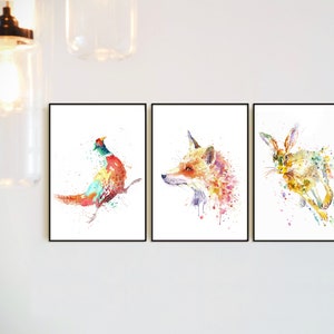 May include: Set of three framed watercolor animal prints. The prints feature a pheasant, a fox, and a hare, each rendered in vibrant, colorful splashes against a white background. The artwork is framed in black.