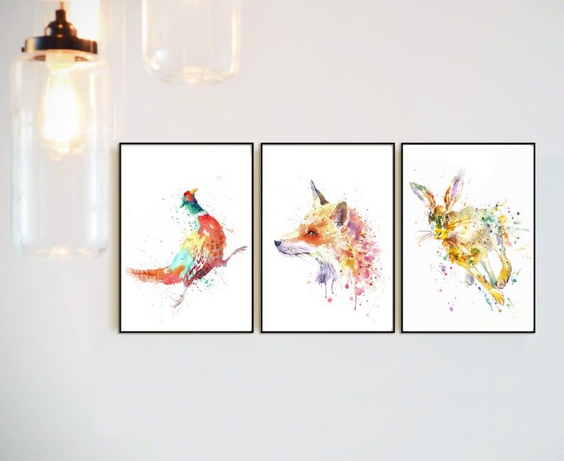 May include: Set of three framed watercolor animal prints. The prints feature a pheasant, a fox, and a hare, each rendered in vibrant, colorful splashes against a white background. The artwork is framed in black.