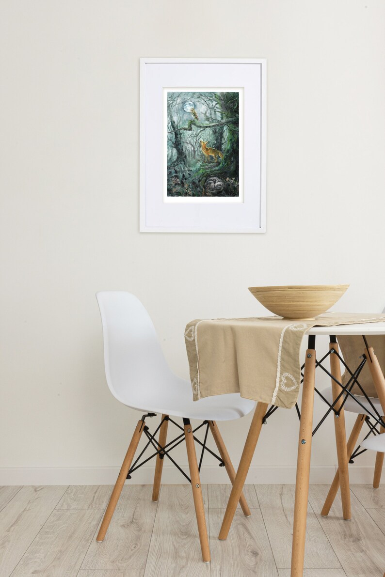 May include: A framed art print depicts a forest scene with a fox, owl, and moon. The artwork is in a white frame and hangs on a white wall. A white chair and table with a wooden bowl and beige tablecloth are in the foreground.