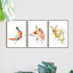 May include: Set of three framed watercolor prints featuring wildlife. The prints depict a pheasant, a fox, and a rabbit, each rendered in a vibrant, colorful, and splattered style. The frames are black.