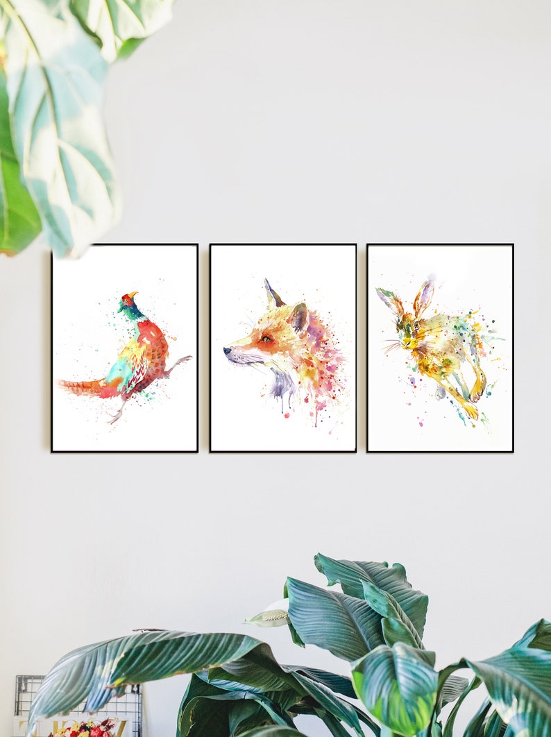 May include: Set of three framed watercolor prints featuring wildlife. The prints depict a pheasant, a fox, and a rabbit, each rendered in a vibrant, colorful, and splattered style. The frames are black.